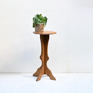 May include: A wooden plant stand with a round top and a decorative, carved base. A small woven basket with a green leafy plant sits on top. The stand is made of light brown wood. The stand is approximately 100 cm tall.