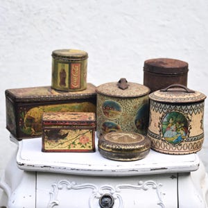 Op de afbeelding: Een verzameling vintage metalen blikken in verschillende vormen en maten, waaronder rechthoekige en cilindrische containers. De blikken hebben verouderde, vervaagde ontwerpen met bloemen, landschappen en Coca-Cola-branding. Ze zijn gerangschikt op een wit oppervlak.