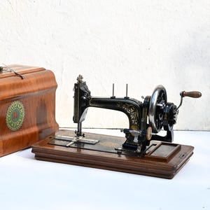 Peut inclure: Une machine à coudre manuelle en métal noir avec des détails floraux dorés, montée sur une base en bois rectangulaire. À côté se trouve une mallette de transport en bois assortie avec une poignée en métal et une étiquette verte Gritzner Extra.
