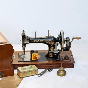Pode incluir: Máquina de costura preta antiga com detalhes florais dourados, sobre uma base de madeira. Acessórios: estojo de madeira, lata pequena e lata de óleo. Um item vintage.