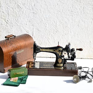 Könnte beinhalten: Eine schwarze Singer-Nähmaschine mit goldenen Filigranverzierungen auf einem Holzsockel mit passender gewölbter Holzabdeckung. Enthält eine grüne Singer-Zubehördose aus Metall, ein kleines grünes Stoffstück mit Blumenstickerei und eine Lampe.