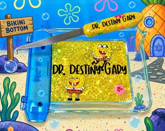 May include: A clear blue plastic tray featuring a gold glitter insert with two SpongeBob SquarePants characters and the text DR. DESTINY GARY. A matching metal tool with a yellow glitter handle labeled DR. DESTINY GARY rests on the tray.