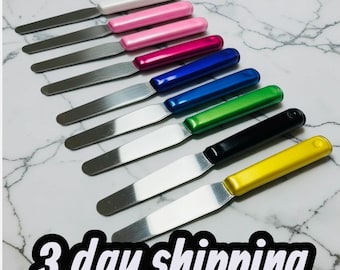May include: An assortment of palette knives with stainless steel blades and colorful handles in white, pink, red, blue, green, black, and yellow. The tools are arranged on a marble-patterned surface.