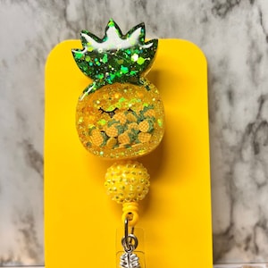 Puede incluir: Un carrete de insignia de piña de brillo amarillo y verde con un pequeño amuleto de piña plateado. El carrete está sobre un fondo amarillo.
