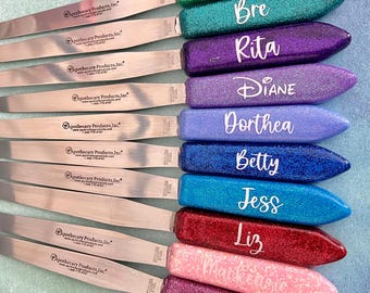 May include: A collection of personalized glitter spatulas in various colors, including green, teal, purple, blue, red, and pink. Each spatula features a different name in white script. The spatulas have stainless steel blades and are labeled with "Apothecary Products, Inc."