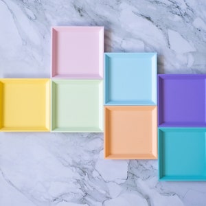 May include: Seven square trays in different pastel colours, arranged in a staggered pattern on a white marble surface. The colours are light pink, light blue, yellow, light green, orange, purple, and turquoise.
