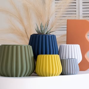May include: Six different coloured ceramic planters with a ribbed design. The planters are arranged in a row on a white surface. The planters are green, blue, white, yellow, and grey.