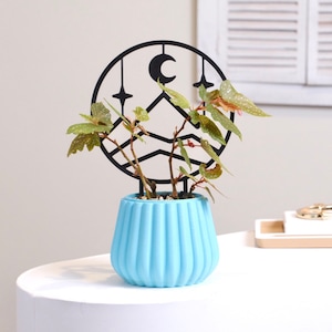May include: A light blue planter with a black metal plant stand featuring a crescent moon, stars, and mountain silhouettes. The planter has a ribbed design and is filled with a green plant.