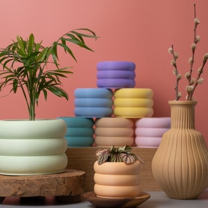 May include: A collection of colourful, textured ceramic planters with a green plant in one and a small plant in another. The planters are stacked in a pyramid shape and are arranged on a wooden surface. A vase with a brown, textured finish holds a sprig of dried branches with fluffy white buds.