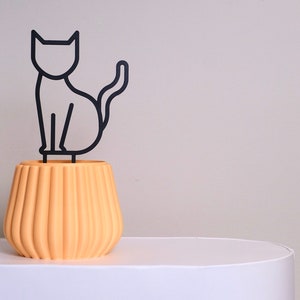 May include: A yellow ceramic planter with a black metal cat silhouette on top. The planter has a ribbed design.