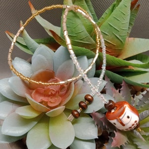 May include: A white and gold beaded necklace with a brown wooden owl charm. The necklace is lying on a green succulent plant.