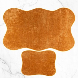 Può includere: Due tappetini da bagno marroni e pelosi con una forma ondulata organica. Il tappetino più grande è sullo sfondo e quello più piccolo è in primo piano.