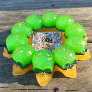 May include: A resin ashtray featuring a ring of green skull shapes with gold flecks, surrounding a central image with the text "King Gizzard & The Lizard Wizard". The ashtray has a yellow base.