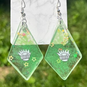 May include: A pair of green diamond-shaped earrings with silver hooks. The earrings have a green glitter background with a white cartoon character with a crown and a mouth with teeth. There are also small yellow stars on the earrings.