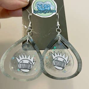 May include: Silver-toned dangle earrings with a teardrop shape. Each earring features a clear center with a cartoon face design and glitter accents, encased in a gray resin frame. The earrings are suspended from silver hooks.