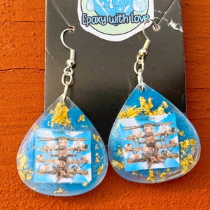 May include: Teardrop-shaped earrings with a blue resin base, gold flakes, and a small image of a structure. The earrings have silver-colored hooks and are displayed against an orange wooden surface.