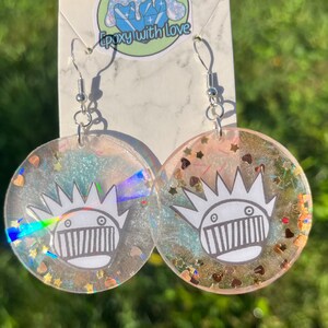 May include: Round, clear resin earrings with a white cartoon face design. The earrings have silver hooks and are filled with iridescent glitter, stars, and heart-shaped confetti. The background is a blurred green.