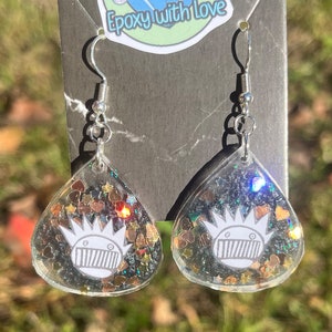May include: Clear resin teardrop earrings with silver hooks. Each earring features a white cartoon face design with a crown and a mouth, surrounded by glitter and heart-shaped confetti. The earrings are displayed against a dark gray background.