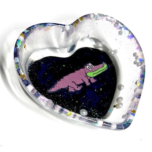 May include: A heart-shaped trinket dish with a clear, glitter-filled rim. Inside, a purple alligator with a green snout is set against a dark blue, starry background. Small, white pearl-like beads are scattered around the edge.