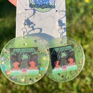 May include: A pair of handmade, round earrings with a green, glittery resin base. Each earring features a small square image with the text "KING GIZZARD & THE LIZARD WIZARD" and a graphic. Silver-colored ear wires complete the design.