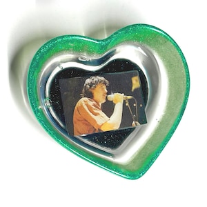 May include: A heart-shaped trinket dish with a green glitter rim and a clear center. Inside, a small rectangular photo shows a person singing into a microphone. The dish is on a white background.