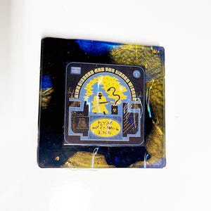 May include: Square coaster with a black, blue, and gold resin design. A smaller square sticker is centered on the coaster, featuring the text "KING GIZZARD AND THE LIZARD WIZARD" and a yellow graphic.