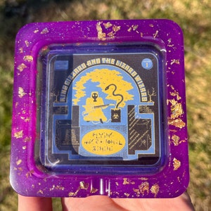 Puede incluir: Un cenicero cuadrado de color morado con purpurina dorada y una imagen de la portada del álbum de King Gizzard and the Lizard Wizard. La portada del álbum muestra una figura con una máscara de gas tocando la guitarra con una serpiente envuelta alrededor de un árbol. El título del álbum es "Flying Microtonal Banana" y el número del álbum es "I".