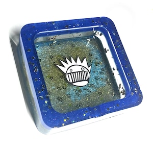 Puede incluir: Bandeja cuadrada de resina con borde de purpurina azul e interior azul y verde. La bandeja presenta un gráfico en blanco y negro en el centro. La bandeja está sobre un fondo blanco.