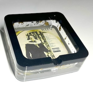 May include: Black ashtray with a clear top, featuring a Ween album cover with a person in black clothing. The ashtray has a speckled, glittery finish.