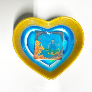 May include: A heart-shaped dish with a yellow rim and a blue interior. The dish contains a square, blue and orange sticker with a figure in the centre.