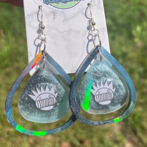 May include: A pair of silver-toned dangle earrings. Each earring features a teardrop-shaped design with a light blue, iridescent interior and a silver-toned outer frame. A white graphic is visible inside the teardrop.