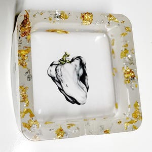 May include: Square ashtray with a clear resin border embedded with gold and silver leaf flakes. The center features a black and white illustration of a bell pepper. A decorative piece for home decor.