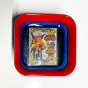 Puede incluir: Una bandeja de resina roja y azul con borde negro, que contiene un cartel de concierto vintage con un personaje de dibujos animados con cabeza roja y cuerpo negro. El texto del cartel dice "King Gizzard and the Lizard Wizard".