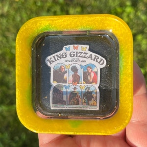 May include: A yellow and green resin tray with a clear glass insert featuring a sticker of the band King Gizzard and the Lizard Wizard. The sticker has a black background with a white border and features the band's name and logo.