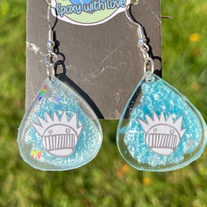 May include: A pair of teardrop-shaped earrings with a light blue, glittery background. Each earring features a white and gray graphic of a crown-like face. The earrings have silver-colored hooks.