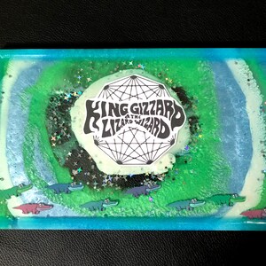 May include: A rectangular resin tray with a turquoise, green, and white swirling design. The tray features the text "KING GIZZARD & THE LIZARD WIZARD" and cartoon alligators. Glitter is sprinkled throughout the resin.