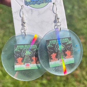 May include: Clear, circular earrings with a silver hook. Each earring features a small, rectangular image with the text "KING GIZZARD & THE LIZARD WIZARD" and a graphic of a green figure. The earrings have a holographic rainbow accent.