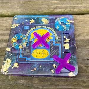 May include: A handmade tic-tac-toe game board with a cosmic theme. The board is a square with a blue and purple galaxy design, gold leaf accents, and the text "Flying Microtonal Banana". The game pieces are purple X's and blue O's.