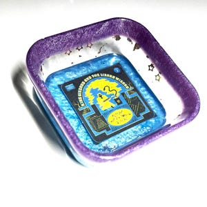 Puede incluir: Un plato cuadrado de resina de color morado y azul con un acabado de purpurina dorada. El plato presenta una imagen impresa de una portada de álbum de disco con el texto "King Gizzard and the Lizard Wizard" y "Fishing for Fishies".