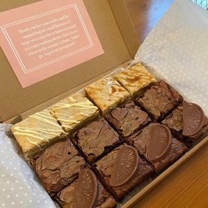 May include: A box filled with a variety of brownies. The brownies are arranged in a grid pattern and include flavors like caramel, white chocolate, and orange. The box is lined with white paper with small polka dots. The box has a thank you note that reads "Thank you for your order and for supporting my small business! I hope you enjoy these bakes as much as I enjoy making them. I love to see where in the country @busy_bakes go - tag us on Instagram!"