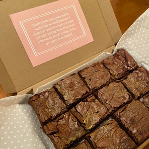 May include: A box filled with a dozen chocolate brownies, each with a swirl of caramel. The box is lined with white paper with a polka dot pattern. A pink card with a thank you message is visible in the background.