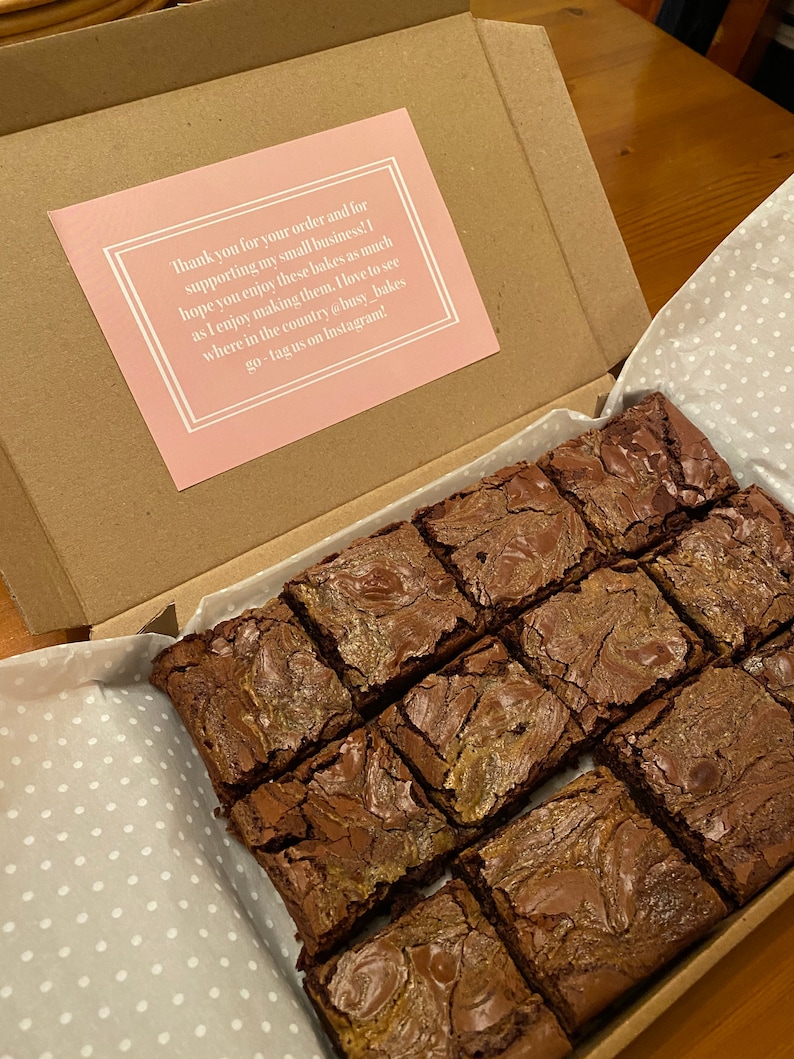 May include: A box filled with a dozen chocolate brownies, each with a swirl of caramel. The box is lined with white paper with a polka dot pattern. A pink card with a thank you message is visible in the background.