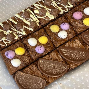May include: A tray of chocolate brownies topped with different decorations. Some brownies have white chocolate drizzled over them with chocolate stars, others have mini chocolate eggs, and some have chocolate orange slices.