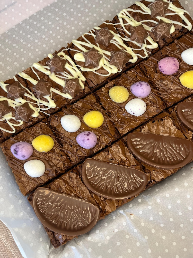 May include: A tray of chocolate brownies topped with different decorations. Some brownies have white chocolate drizzled over them with chocolate stars, others have mini chocolate eggs, and some have chocolate orange slices.