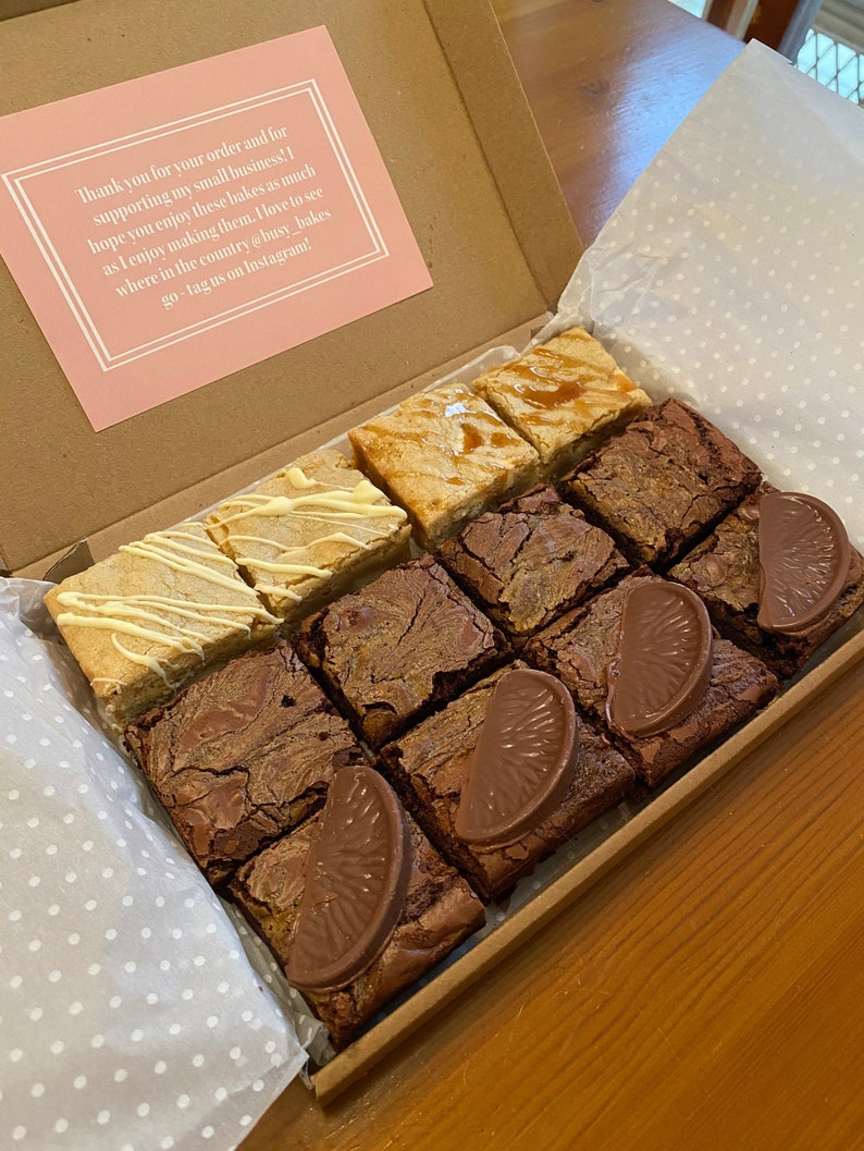 May include: A box filled with a variety of brownies. The brownies are arranged in a grid pattern and include flavors like caramel, white chocolate, and orange. The box is lined with white paper with small polka dots. The box has a thank you note that reads "Thank you for your order and for supporting my small business! I hope you enjoy these bakes as much as I enjoy making them. I love to see where in the country @busy_bakes go - tag us on Instagram!"