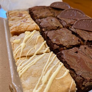 May include: A box of assorted brownies and blondies. The brownies are dark chocolate and have a marbled appearance. The blondies are topped with white chocolate drizzle.
