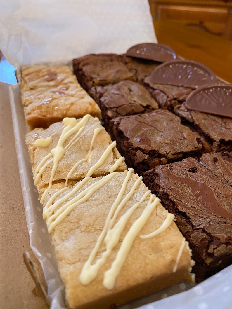 May include: A box of assorted brownies and blondies. The brownies are dark chocolate and have a marbled appearance. The blondies are topped with white chocolate drizzle.