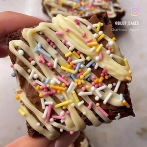 May include: A close-up of a chocolate brownie topped with white chocolate and colorful sprinkles. The brownie is being held by a hand. The text "@busy_bakes Charli xcx + Apple" is visible in the image.