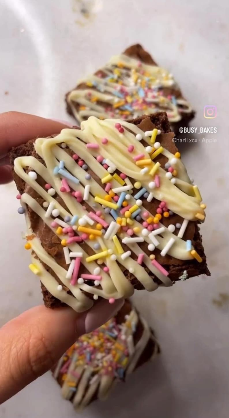May include: A close-up of a chocolate brownie topped with white chocolate and colorful sprinkles. The brownie is being held by a hand. The text "@busy_bakes Charli xcx + Apple" is visible in the image.