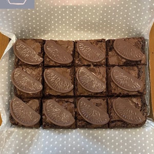 May include: A box of 16 chocolate brownies with orange slices on top. The brownies are arranged in a 4x4 grid. The box is lined with white paper with a polka dot pattern.
